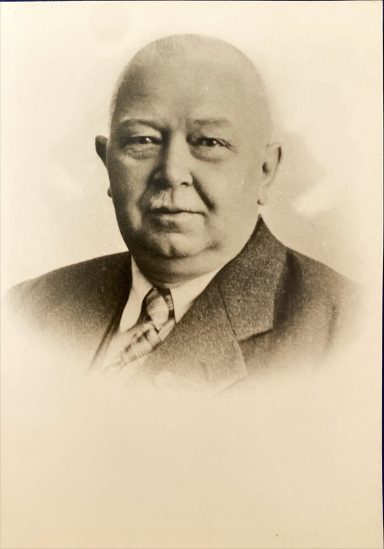 Porträt eines Mannes in Anzug mit neutralem Gesichtsausdruck, sepiafarben.